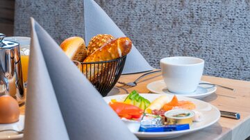 A laid breakfast table with fresh rolls, a cup of coffee, and fresh fruit. In the background, decorated napkins can be seen.