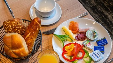 A breakfast table with fresh rolls, cold cuts, cheese, and colorful vegetables. Alongside is a glass of orange juice and an empty cup.