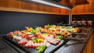 A rich buffet with various sausage and cheese plates, garnished with fresh vegetables and fruits. The presentation is appealingly arranged on a dark table.