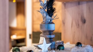 A stylish decoration with a silver vase, black and green balls, and a white fur base. In the background, a cozy room is visible.