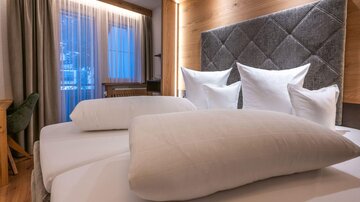 A cozy bedroom with a large bed and many pillows. The design is modern and inviting, with warm wood tones and natural light.