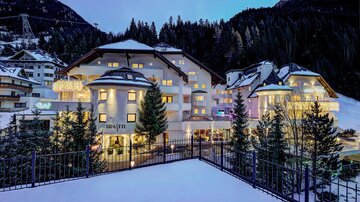 An elegant hotel in the Alps, surrounded by snow-covered trees. The facade glows softly in the evening light.