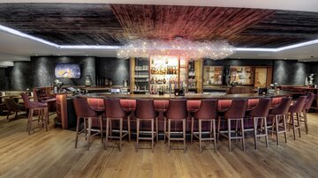 A modern bar with a curved counter area and stylish, ultra-modern bar stools. The space has a warm atmosphere with wooden flooring and indirect lighting.