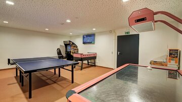 A room with a ping pong table and an air hockey table. In the background, there is a television, and there are other gaming devices.