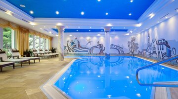 A modern indoor swimming pool with clear blue water. On the walls are decorative murals and the sky is in a bright blue shade.