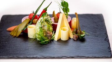 An artistically arranged plate with various vegetables, cheese, and small side dishes. The presentation is on a black plate.
