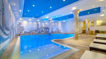 A modern indoor pool with bright blue water. The walls are adorned with elaborate murals, and the ceiling light creates a pleasant atmosphere.