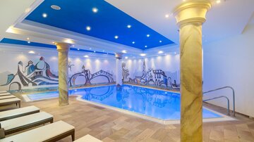An elegant indoor pool with a blue sky above the water. Surrounded by comfortable loungers and decorative walls.