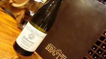 A bottle of wine with a white label stands on a wooden table. In the background, a menu or wine list can be seen.