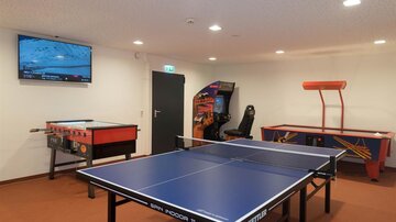 A recreational area with a table tennis table, a foosball table, and a pinball machine. In the background, there is a television on the wall.