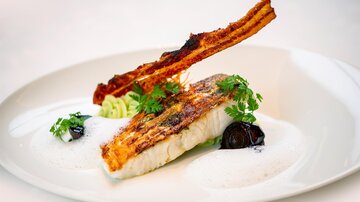 A finely arranged plate with fried fish, garnished with crispy bacon and fresh herbs. Accompanied by a frothy sauce and black olives.