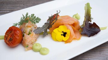 An attractively arranged dish with salmon, a fried tomato, and fresh herbs. Decorative flowers and green sauces provide a colorful accent.