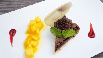 A piece of chocolate cake with a decorative whipped cream topping and a mint leaf. Next to the cake are fresh mango cubes and fruit sauce.