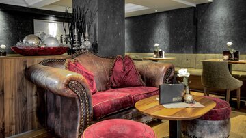 A stylish lounge with a red velvet sofa and a round table. The walls are dark and subtly decorated, creating a cozy atmosphere.