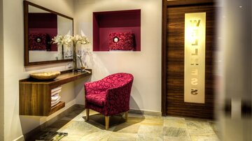 An elegant reception area with a red chair and a mirror. A stylish wooden table with decorations and a labeled access.