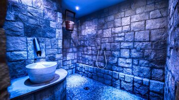 A modern shower area with stone walls and blue light accents. A round washbasin is located next to a showerhead.