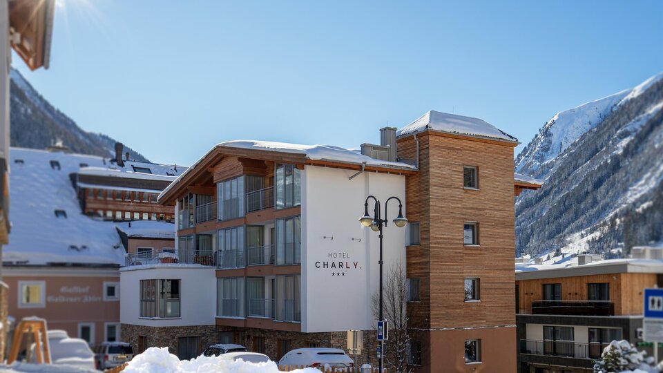 A modern hotel building with a wooden facade and large windows, surrounded by snow-covered mountains. The sky is clear and blue, creating an inviting atmosphere.