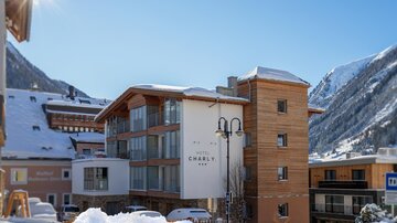 A modern hotel building with a wooden facade and large windows, surrounded by snow-covered mountains. The sky is clear and blue, creating an inviting atmosphere.