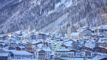 A picturesque winter town with snow-covered houses. In the background, a wooded hill rises under a clear sky.