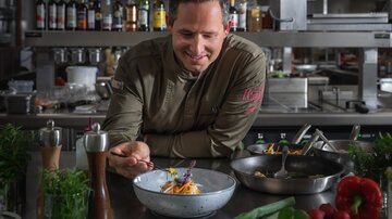 A chef in a modern kitchen proudly presents a plate with creatively arranged dishes. In the background, cooking utensils and ingredients are visible.