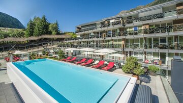 A modern hotel with a large outdoor pool and red loungers. In the background, green mountains and a cozy terrace can be seen.