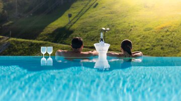A couple relaxes in an infinity pool overlooking the green meadows. Next to them are two glasses and a bottle, ready for a special moment.