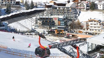 A picturesque ski landscape with snow-covered slopes and a modern gondola lift. In the background, there are mountain hotels and spruces.