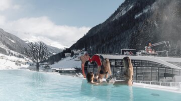 A heated outdoor pool in the mountains, surrounded by snow-covered peaks. Three women enjoy the water and the view in a relaxed atmosphere.