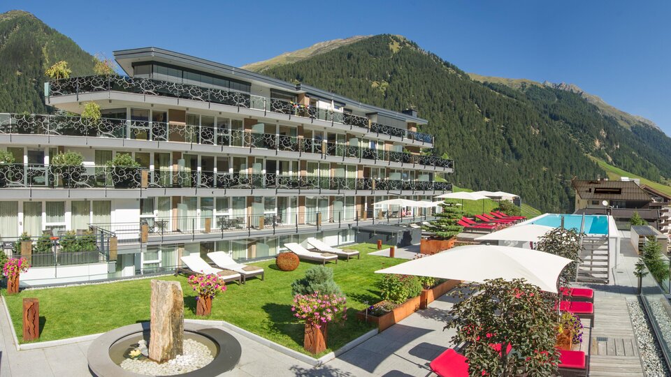 A modern hotel building with balconies and a pool in a beautiful mountain landscape. The outdoor area is designed with loungers and plants.
