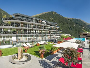 A modern hotel building with balconies and a pool in a beautiful mountain landscape. The outdoor area is designed with loungers and plants.