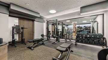 A modern gym with various training machines and weights. The walls are bright and mirrors reflect the room.