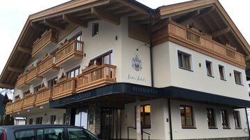 A modern building in alpine style with wooden balconies. Below is a restaurant and a parking lot.