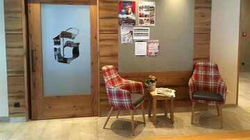 A cozy entrance area with wooden paneling and two checkered chairs. Information sheets are hanging on the wall, and a glass door leads to another room.