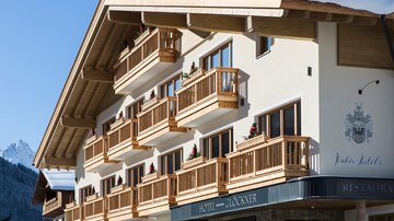 A charming hotel with wooden balconies and a modern alpine design. The surroundings feature snow-covered mountains in the background.