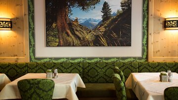 A cozy restaurant with green, patterned benches and tables. A large picture of a mountain landscape hangs on the wall.