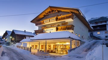 A charming hotel in the snow with an inviting entrance. The mountains are visible in the background and the sky is clear and blue.
