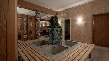 A cozy wellness area with a central fountain and wooden bench. The walls are in warm earthy tones, and plants create a relaxed atmosphere.
