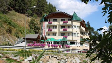 A charming hotel with colorful flower balconies, surrounded by trees. In the background, a bright blue sky can be seen.