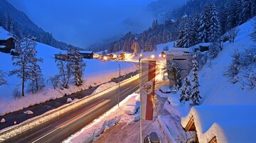 A snowy landscape with a winding path and illuminated vehicles. In the background, snow-covered trees and mountains are visible.