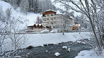 A picturesque winter scene with snow-covered trees and a clear river. In the background, a cozy hotel can be seen, surrounded by the snowy landscape.