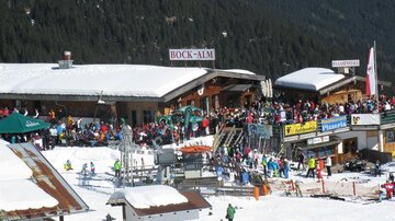 A bustling ski slope with many people. In the foreground, ski huts and stalls are visible in a snow-covered landscape.