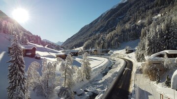A picturesque winter landscape with snow-covered trees and houses. The road winds through the snowy scenery under a bright blue sky.