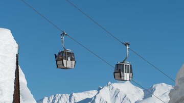 Two gondola lifts glide over snow-covered mountains under a radiant blue sky. The scene conveys a sense of freedom and adventure in the mountains.