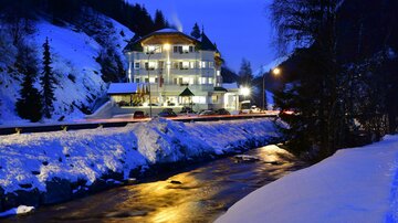 A picturesque hotel in the mountains, surrounded by snow and a river. The night lighting creates a cozy atmosphere.