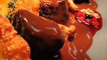An elegant dish with juicy meat, dark sauce, and side dishes. Fresh herbs and a crispy component are also visible.
