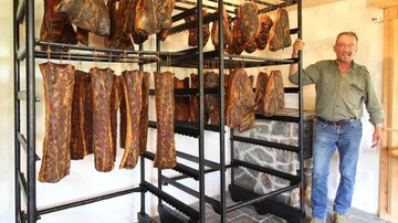 A man stands next to a rack with dried meat. In the background, several pieces of dried meat are hanging in a rural setting.