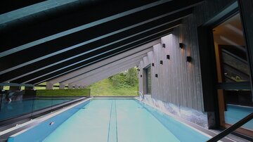 A modern pool under a slanted roof with wooden cladding. The view shows green countryside through large windows.