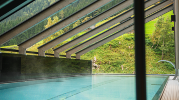 A modern pool area with a glass ceiling. In the background, green hills and mountains can be seen.