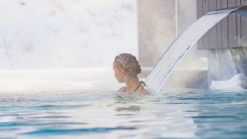 A person is relaxing in the water of an outdoor pool. In the background, snow-covered landscapes and buildings can be seen.