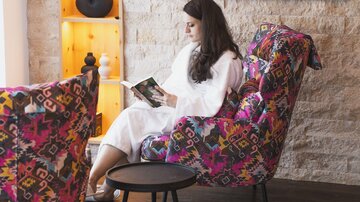A person in a white bathrobe sits relaxed in a colorful armchair and reads a book. In the background, a television and a decorative wall with shelves can be seen.
